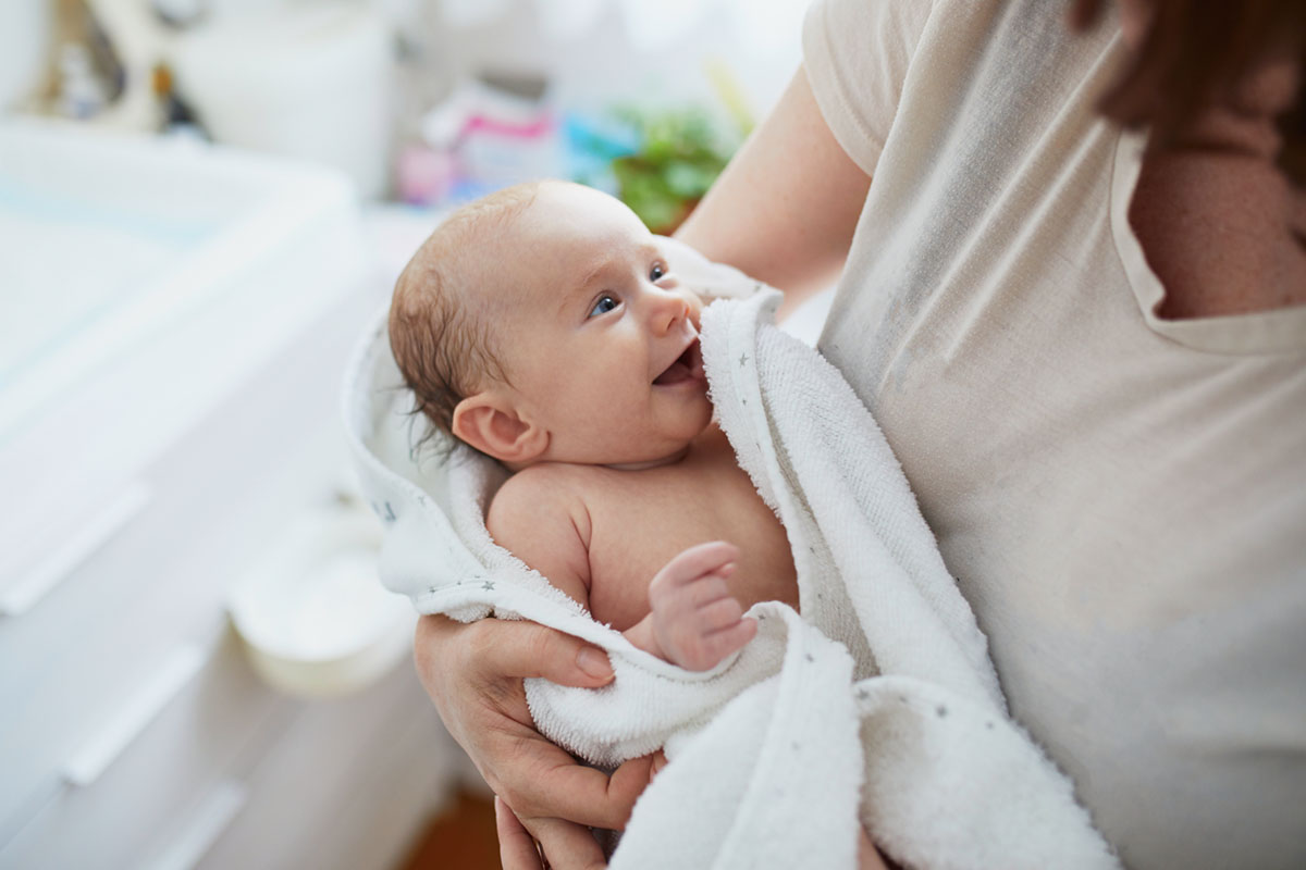 Mutter hält ihr in ein Handtuch gewickeltes Baby nach dem Baden liebevoll auf dem Arm als Sinnbild für das schonende Baden bei trockener Babyhaut
